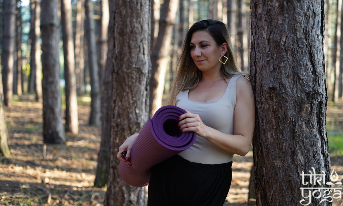 devojka stoji naslonjena na drvo sa protirkom za jogu u prirodi